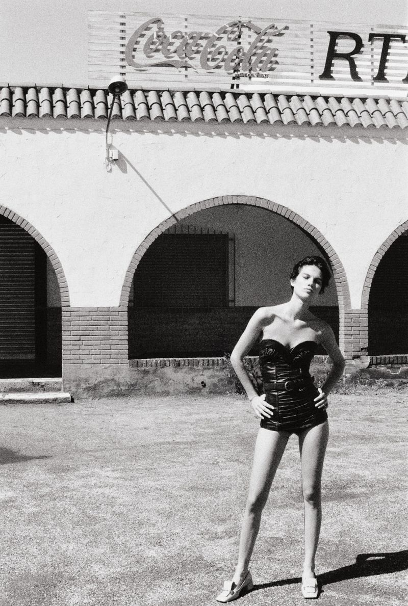 A female fashion model poses in a black swimsuit against an exterior wall with a Coca-Cola sign, captured in a full shot.