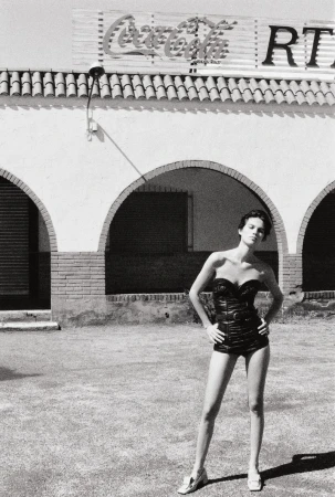 A female fashion model poses in a black swimsuit against an exterior wall with a Coca-Cola sign, captured in a full shot.