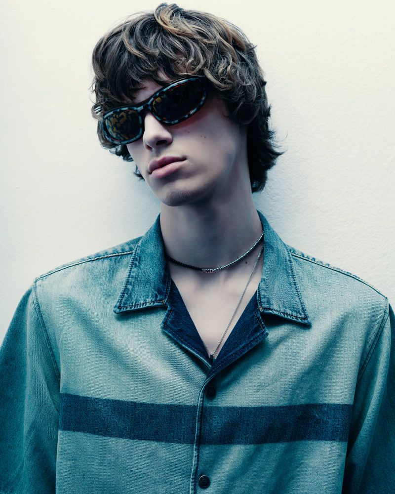 A young man with brown hair and sunglasses is posing indoors. He is wearing a blue denim shirt with a dark blue horizontal stripe.