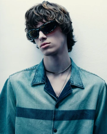 A young man with brown hair and sunglasses is posing indoors. He is wearing a blue denim shirt with a dark blue horizontal stripe.