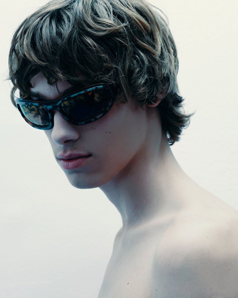 Close-up of a young adult male wearing sunglasses, showcasing wavy brown hair and a white backdrop, portrait orientation.