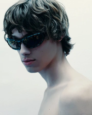 Close-up of a young adult male wearing sunglasses, showcasing wavy brown hair and a white backdrop, portrait orientation.