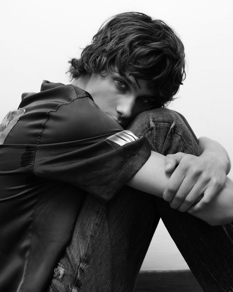 Black and white portrait of a young adult male with dark curly hair, wearing jeans and a short-sleeved shirt, hugging his knees indoors.
