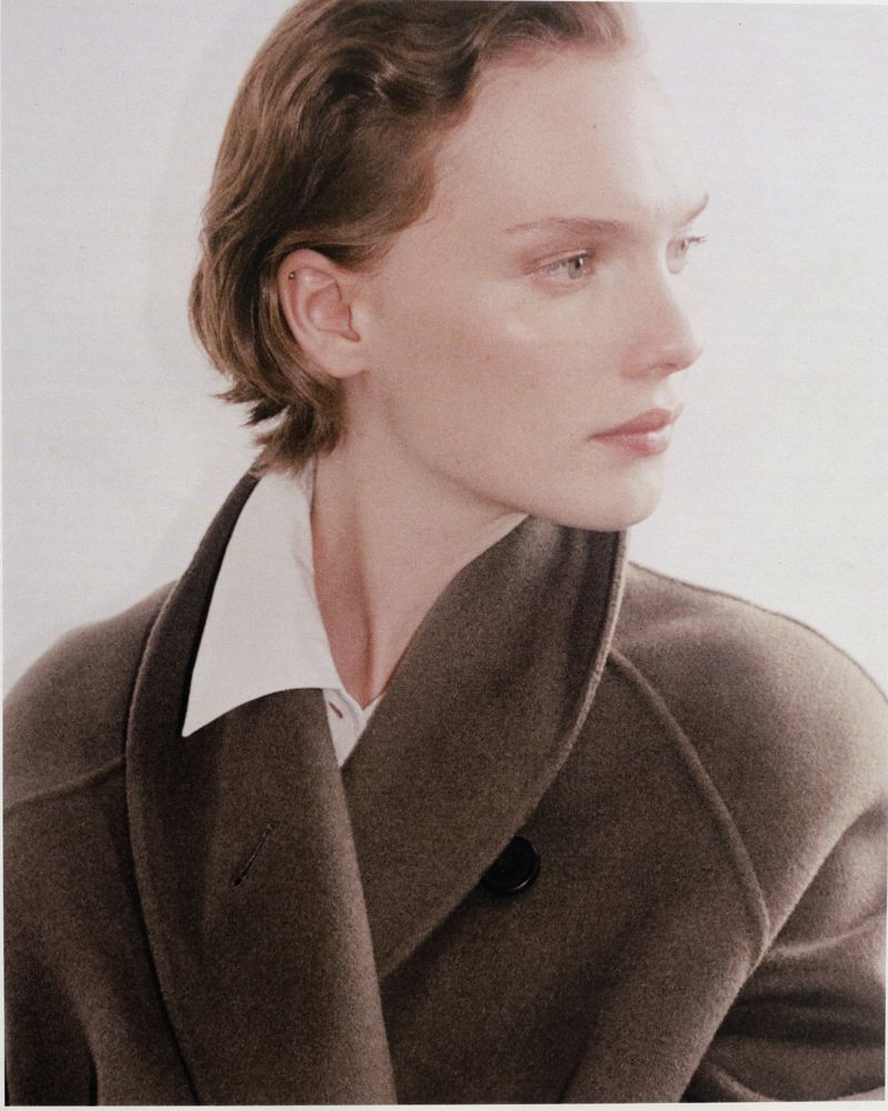 Close-up portrait of an adult woman with short wavy hair wearing a taupe coat over a white collared shirt against a light background.
