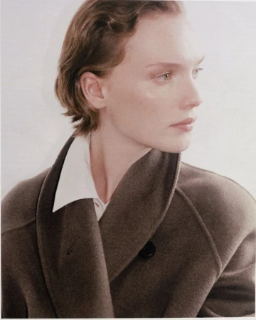 Close-up portrait of an adult woman with short wavy hair wearing a taupe coat over a white collared shirt against a light background.