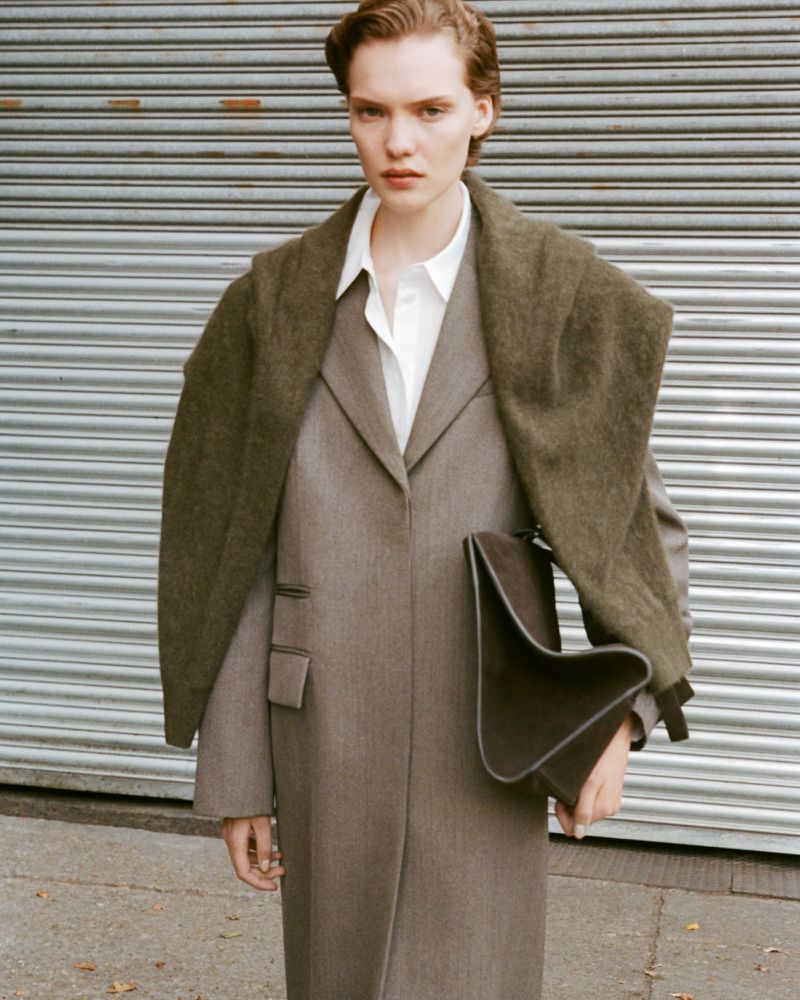 Fashionable adult woman in a gray coat and white shirt carrying a large brown clutch bag, posing in front of metal garage door.