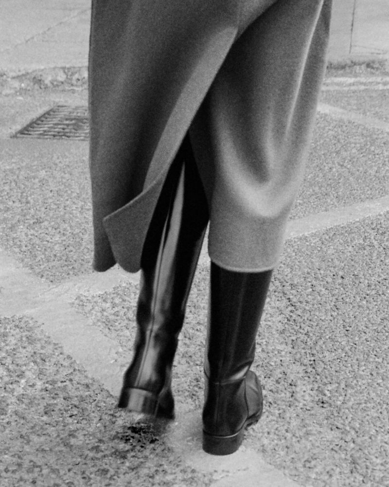 Person wearing a grey coat and black riding boots walks on a paved surface with recessed drain, captured in a grayscale fashion shot.