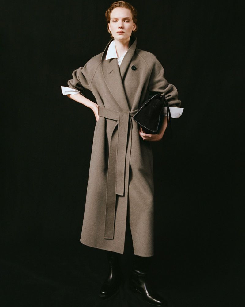 Fashionable woman in a tan wool coat and black boots poses in a photo studio, holding a black leather handbag against a dark backdrop.