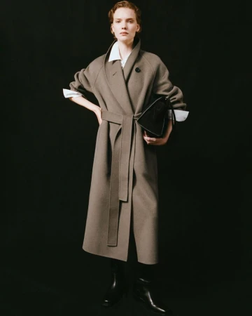 Fashionable woman in a tan wool coat and black boots poses in a photo studio, holding a black leather handbag against a dark backdrop.