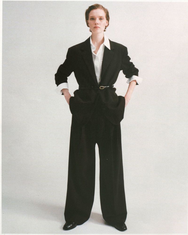 Model wearing an oversized black tailored suit with wide-leg trousers and a white collared shirt against a white studio backdrop.
