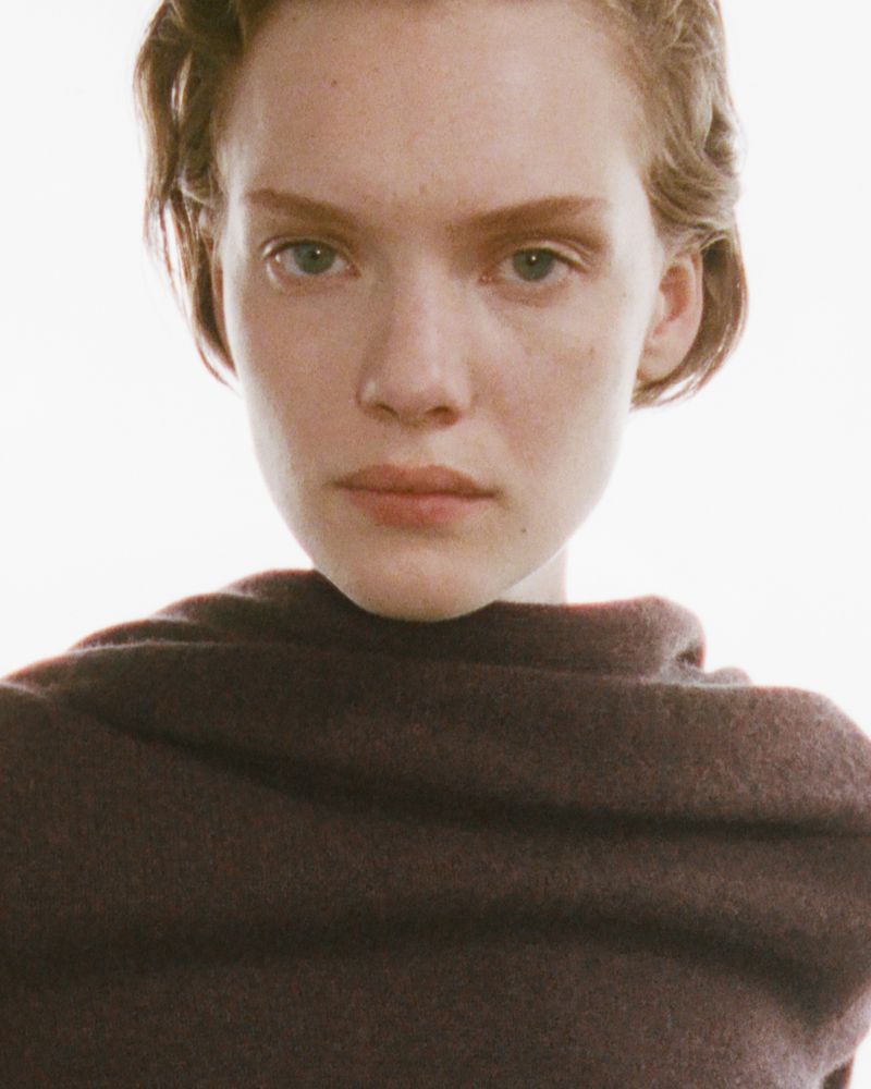 Close-up portrait of a woman with short hair wearing a high-necked top, set against a bright, neutral backdrop in a fashion editorial style.
