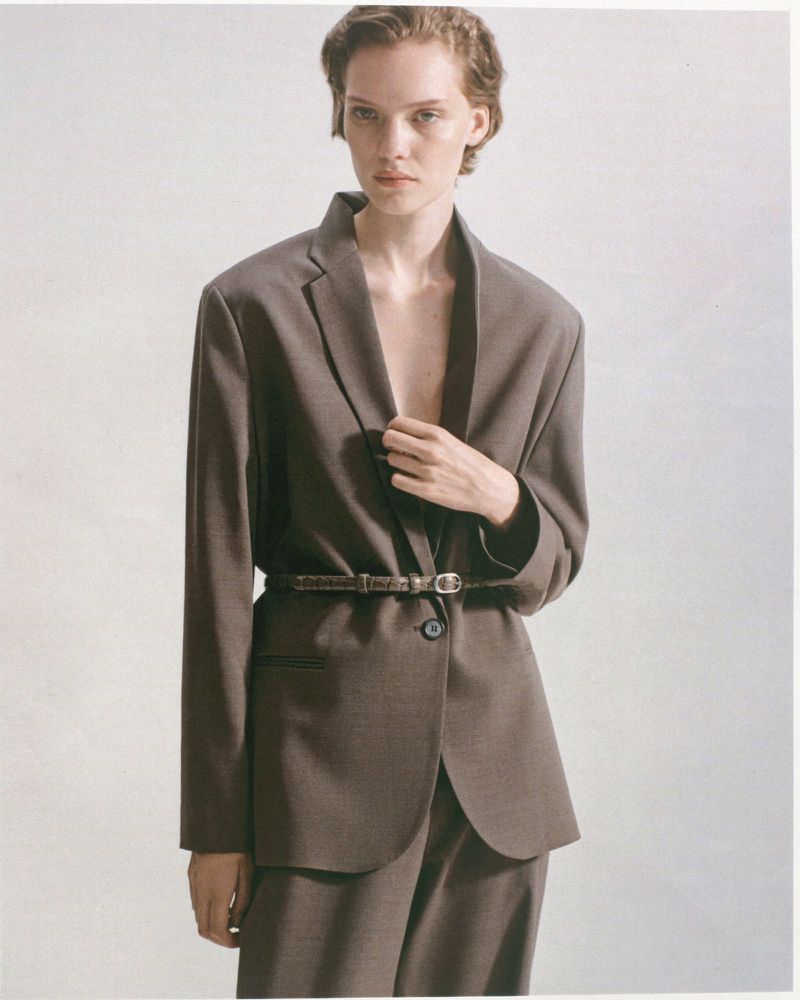 A woman in a brown blazer and trousers stands in a studio. The blazer is cinched at the waist with a thin brown belt. She poses against a white background.