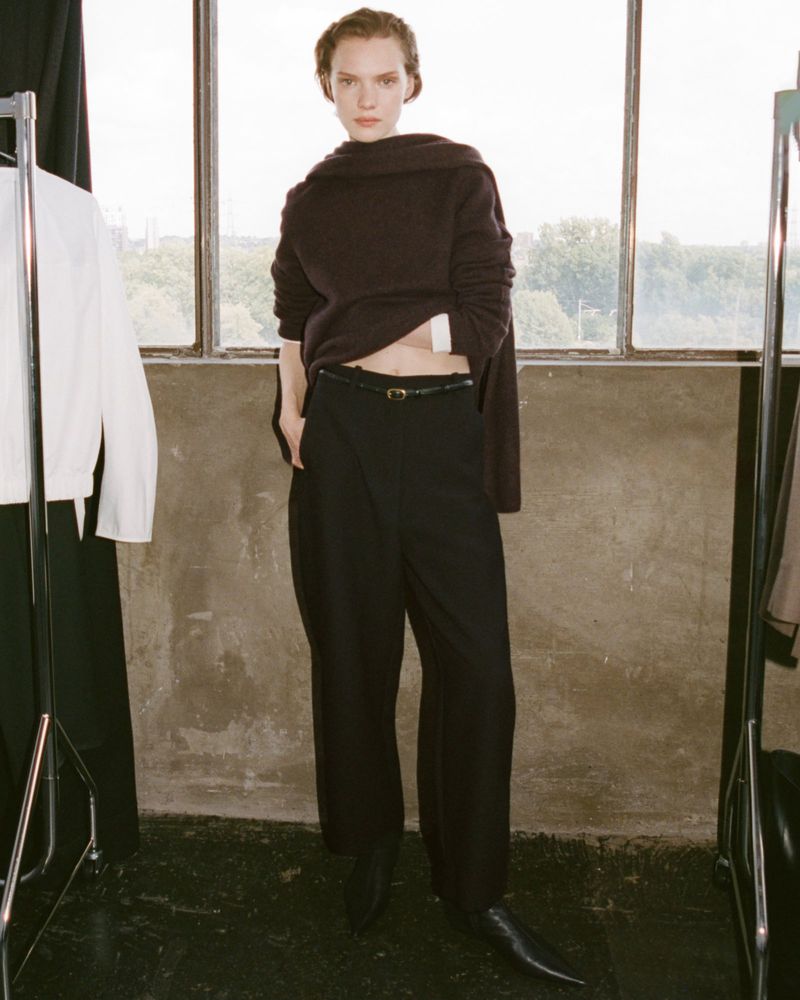 Fashion editorial shot of a model in black wide-leg trousers and a brown sweater, posing by a large window with a city view.