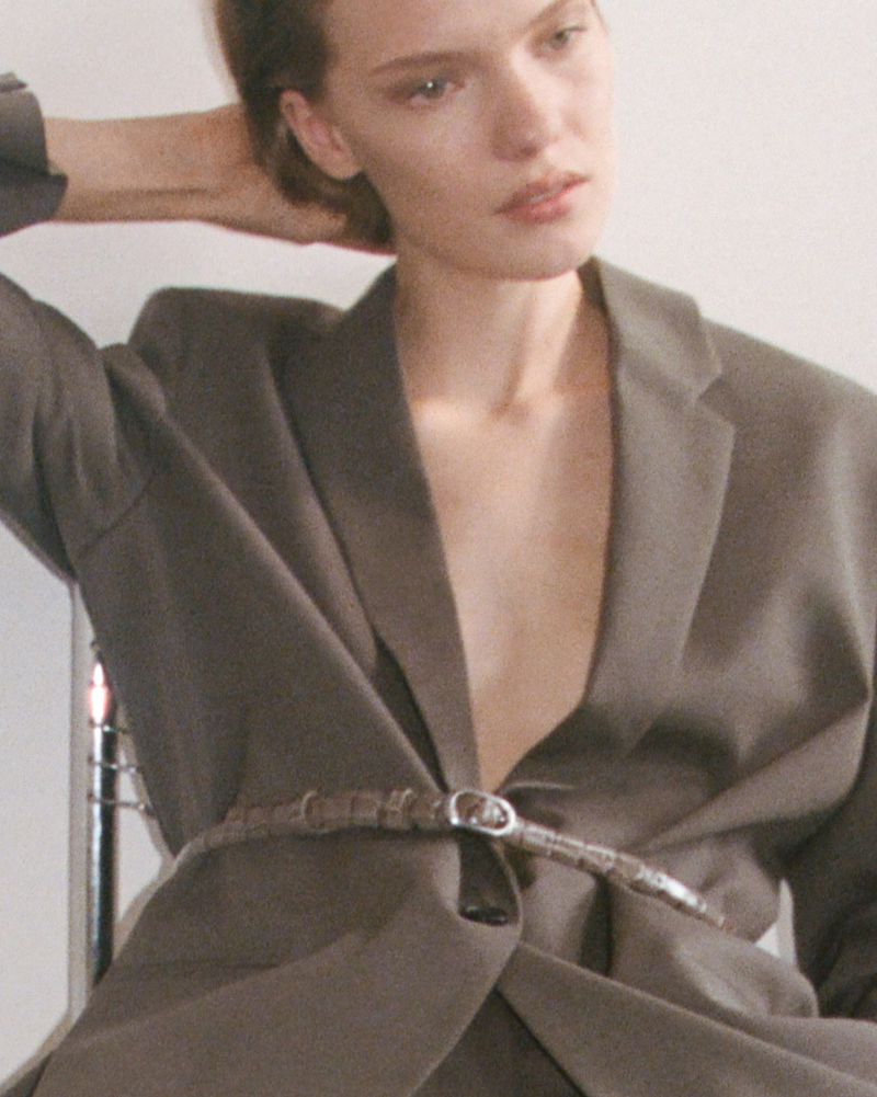 Close-up studio shot of a fair-skinned adult woman wearing a gray blazer and a unique belt, posing against a neutral background.