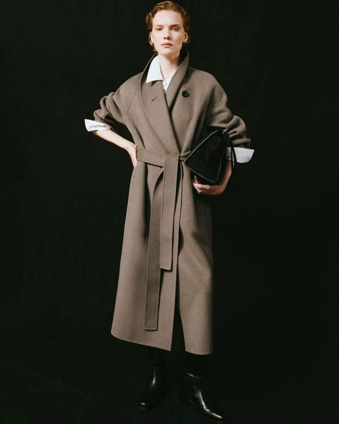 Model Penelope Ternes wearing an oversized belted taupe wool coat and black boots against a solid black background.