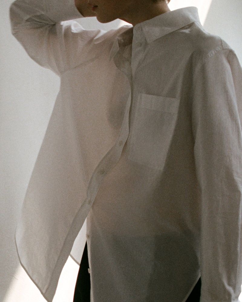 Model wearing a white button-down shirt with chest pocket against a plain wall with natural window light