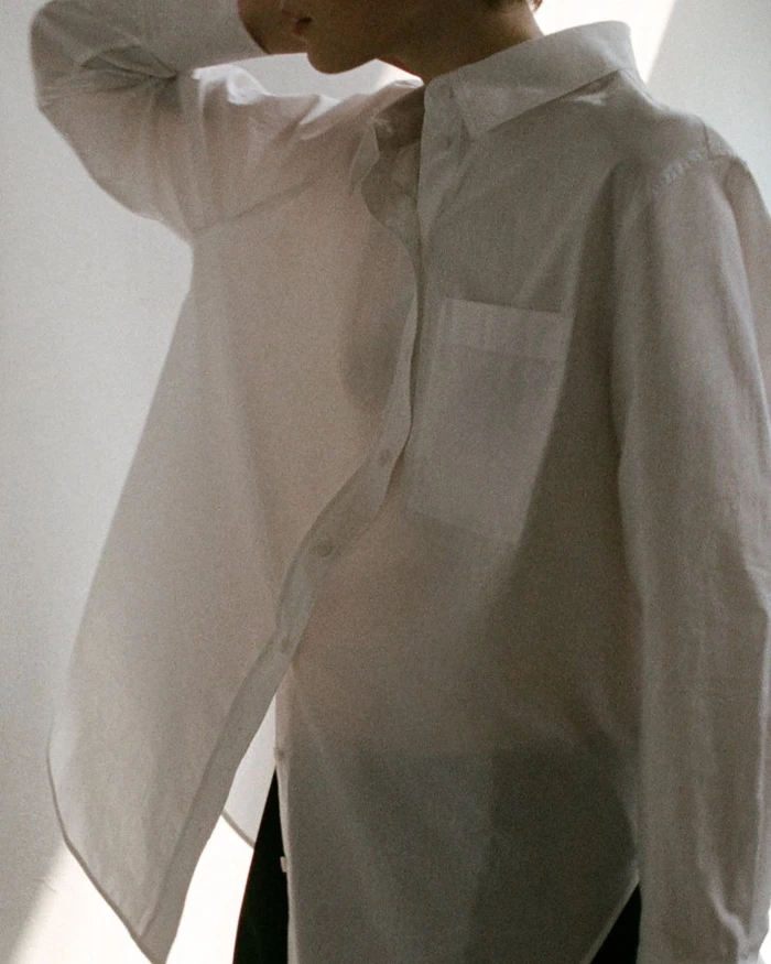 Model wearing a white button-down shirt with chest pocket against a plain wall with natural window light