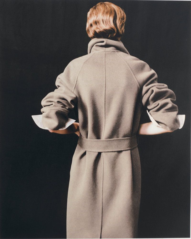 Model Penelope Ternes seen from behind wearing a belted taupe wool coat with white turned-up cuffs against a solid black background.