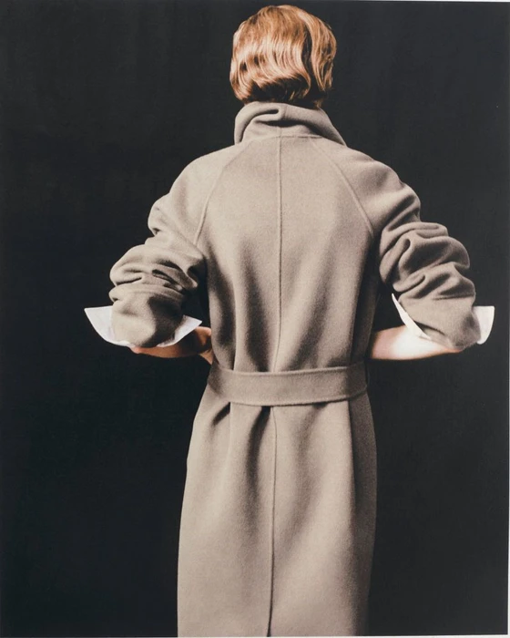 Model Penelope Ternes seen from behind wearing a belted taupe wool coat with white turned-up cuffs against a solid black background.