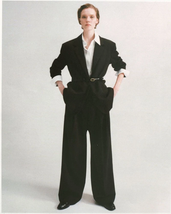 Model wearing an oversized black tailored suit with wide-leg trousers and a white collared shirt against a white studio backdrop.