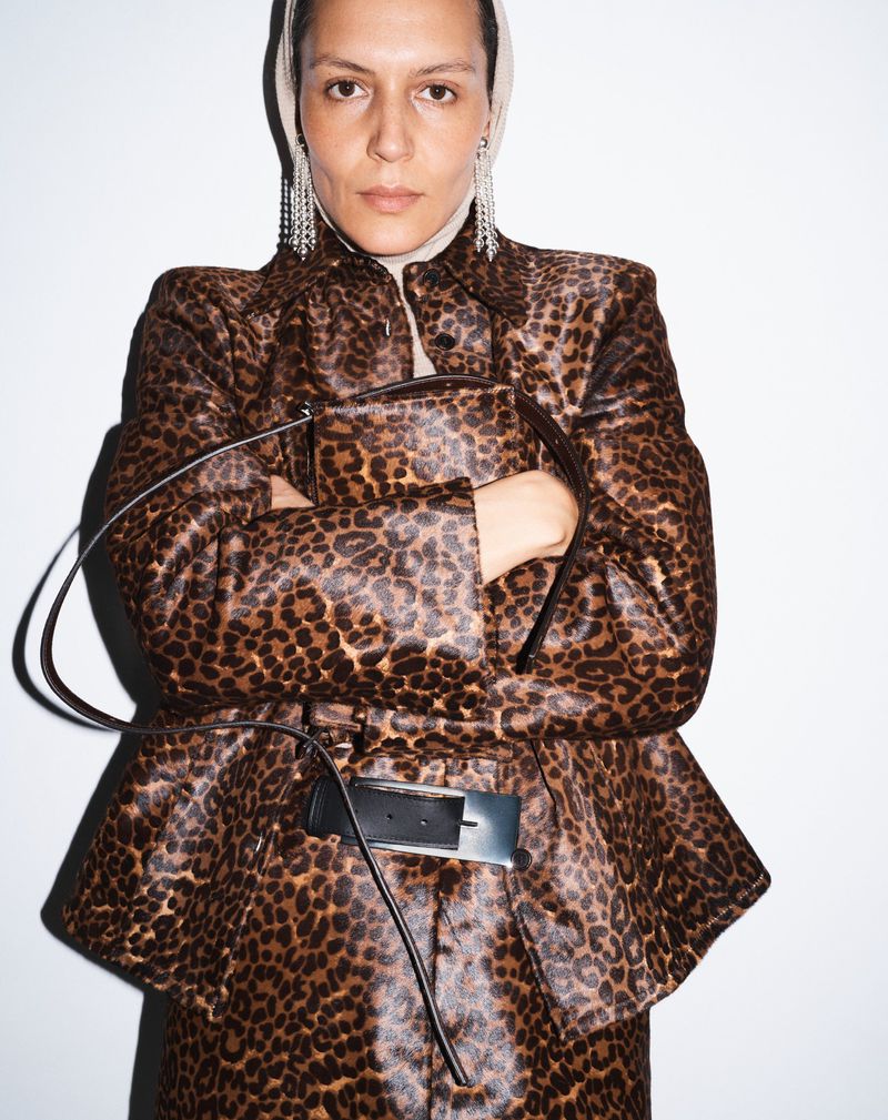 Woman in a leopard-print jacket poses indoors against a white wall, wearing earrings and a head covering, holding a matching purse.