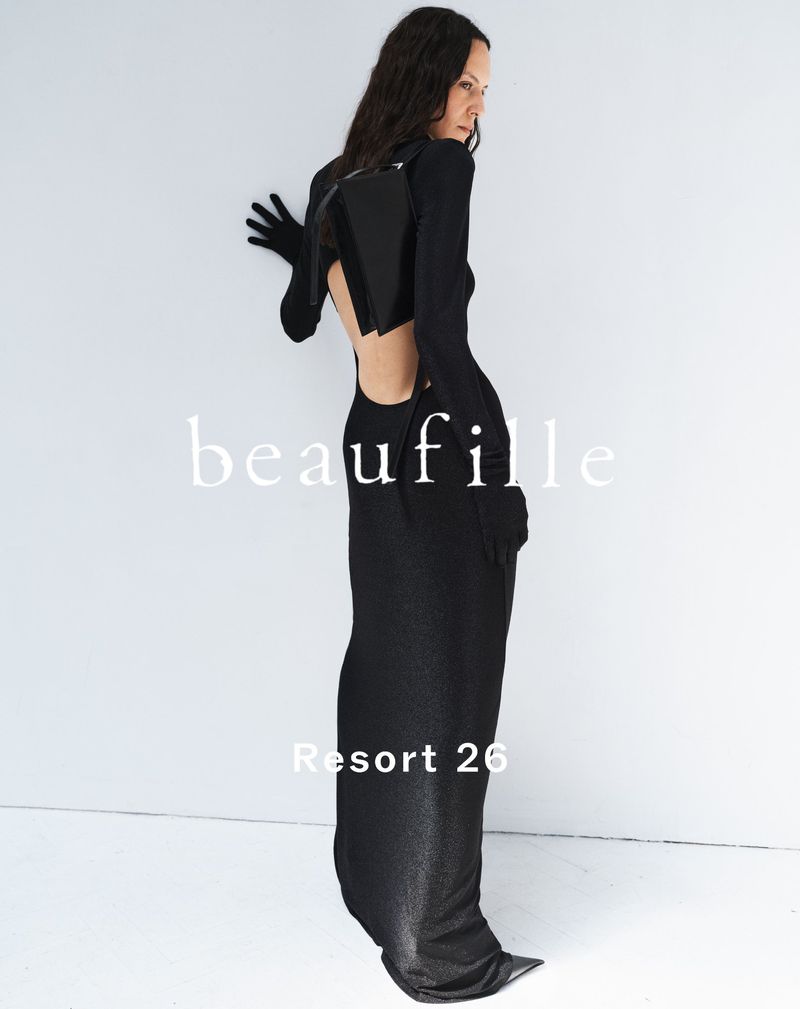 A woman in a full-length black dress poses against a white wall, with view of the back and long sleeves, for a fashion shot.