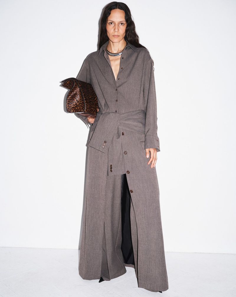 An adult woman poses in a grey button-down outfit with an abstract patterned handbag in a studio setting on a white background.