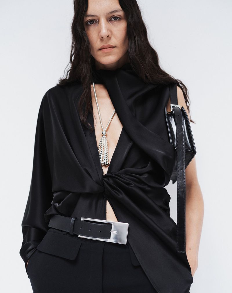 Elegant woman in a black twisted satin top with a silver belt buckle, wearing a necklace, against a plain white backdrop.
