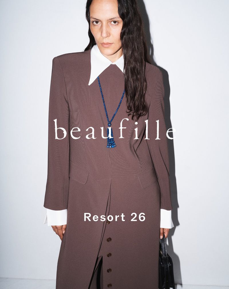 A person poses in a studio wearing a long brown coat with a white collared shirt, necklace, and black purse, against a neutral backdrop.