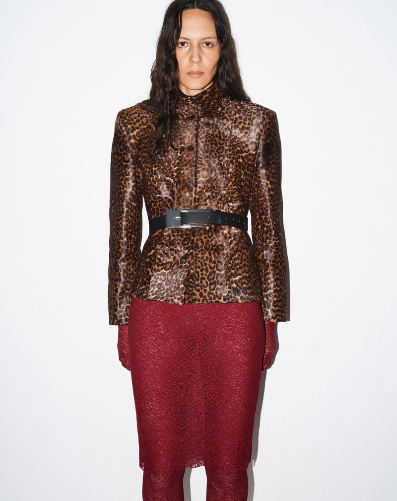 Fashion shot of a woman wearing a leopard print jacket, black belt, and red lace skirt, posing against a white background.