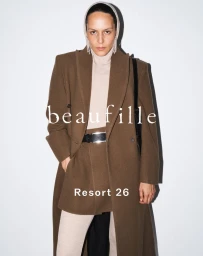 Model Laura Rylands wearing a tailored brown coat, mini skirt, and cream turtleneck against a white background for Beaufille Resort 26.