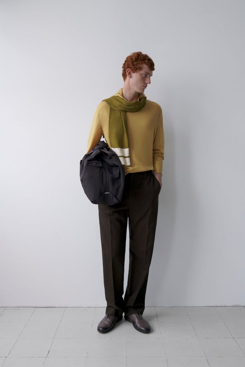 Full-length shot of a red-haired adult male wearing a yellow sweater, green scarf, and brown trousers, holding a black backpack against a white wall.