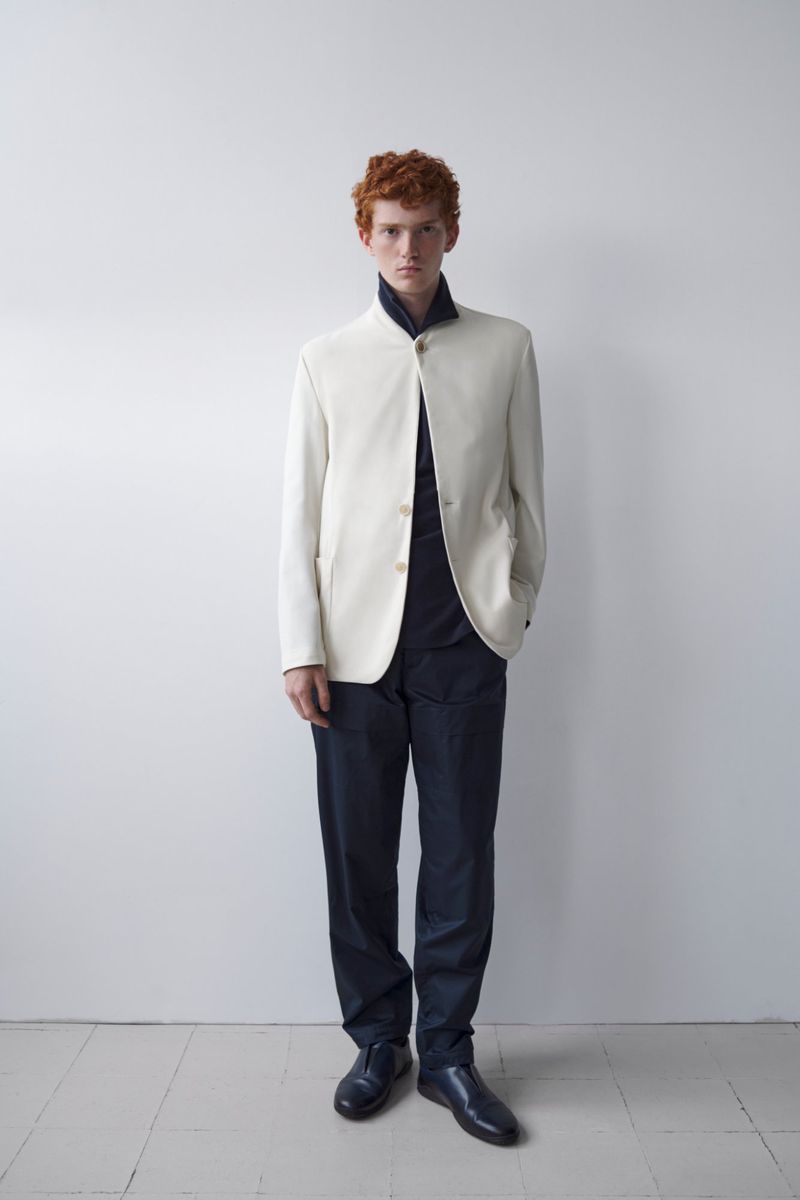 Fashion portrait of a red-haired male model in a cream blazer and navy pants posing in front of a white wall with a neutral expression.
