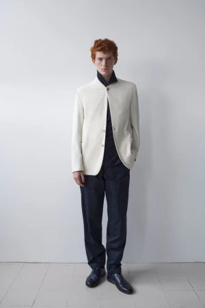 Fashion portrait of a red-haired male model in a cream blazer and navy pants posing in front of a white wall with a neutral expression.