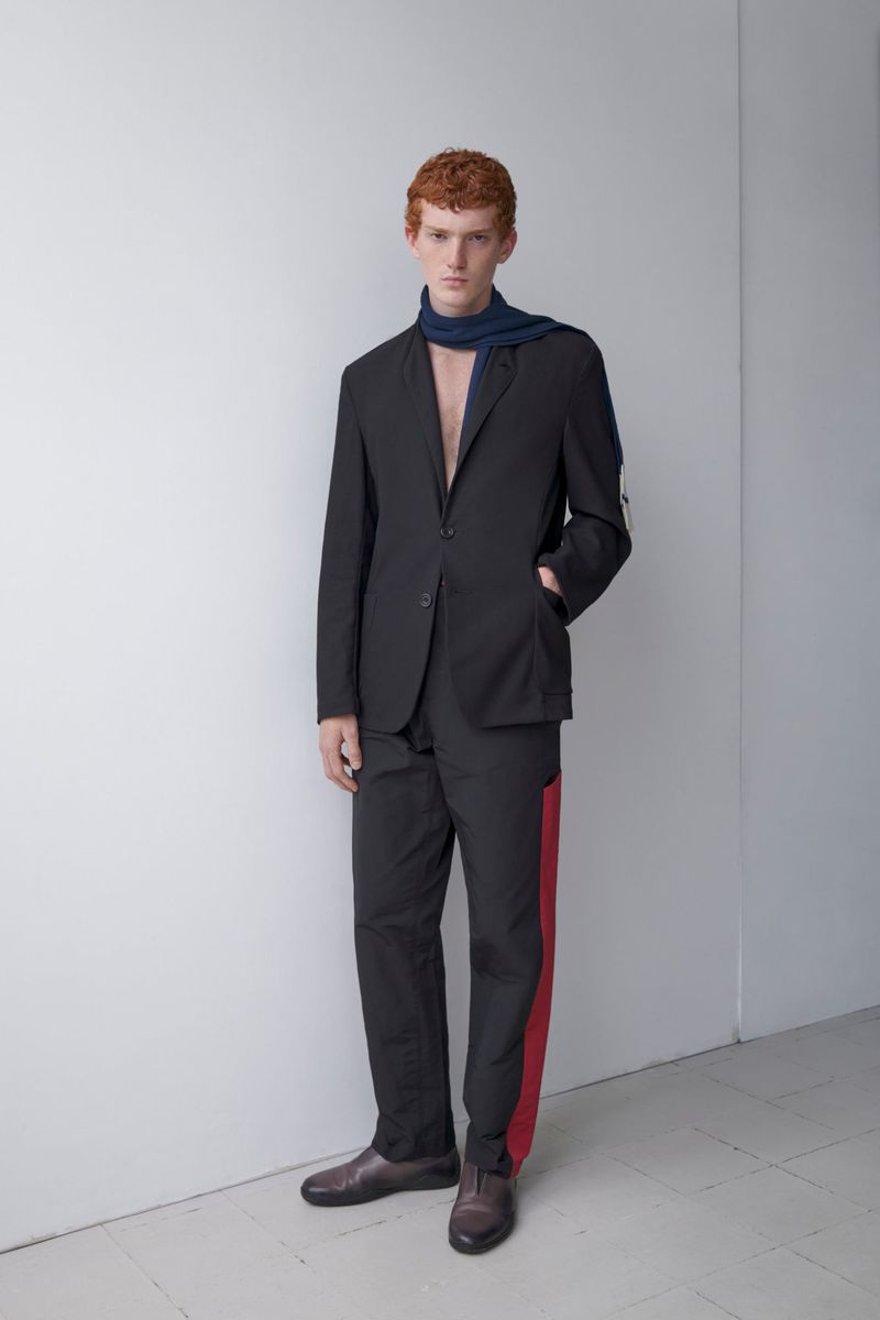 Full shot of a red-haired man wearing a black suit and blue scarf, standing in a minimalist interior with a white wall and brown leather shoes.
