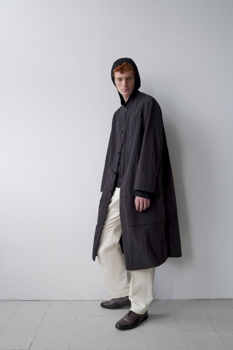 A red-haired male model poses in a long, dark-grey coat with a hood over his head, wearing off-white pants and dark shoes in a studio setting.