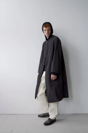 A red-haired male model poses in a long, dark-grey coat with a hood over his head, wearing off-white pants and dark shoes in a studio setting.