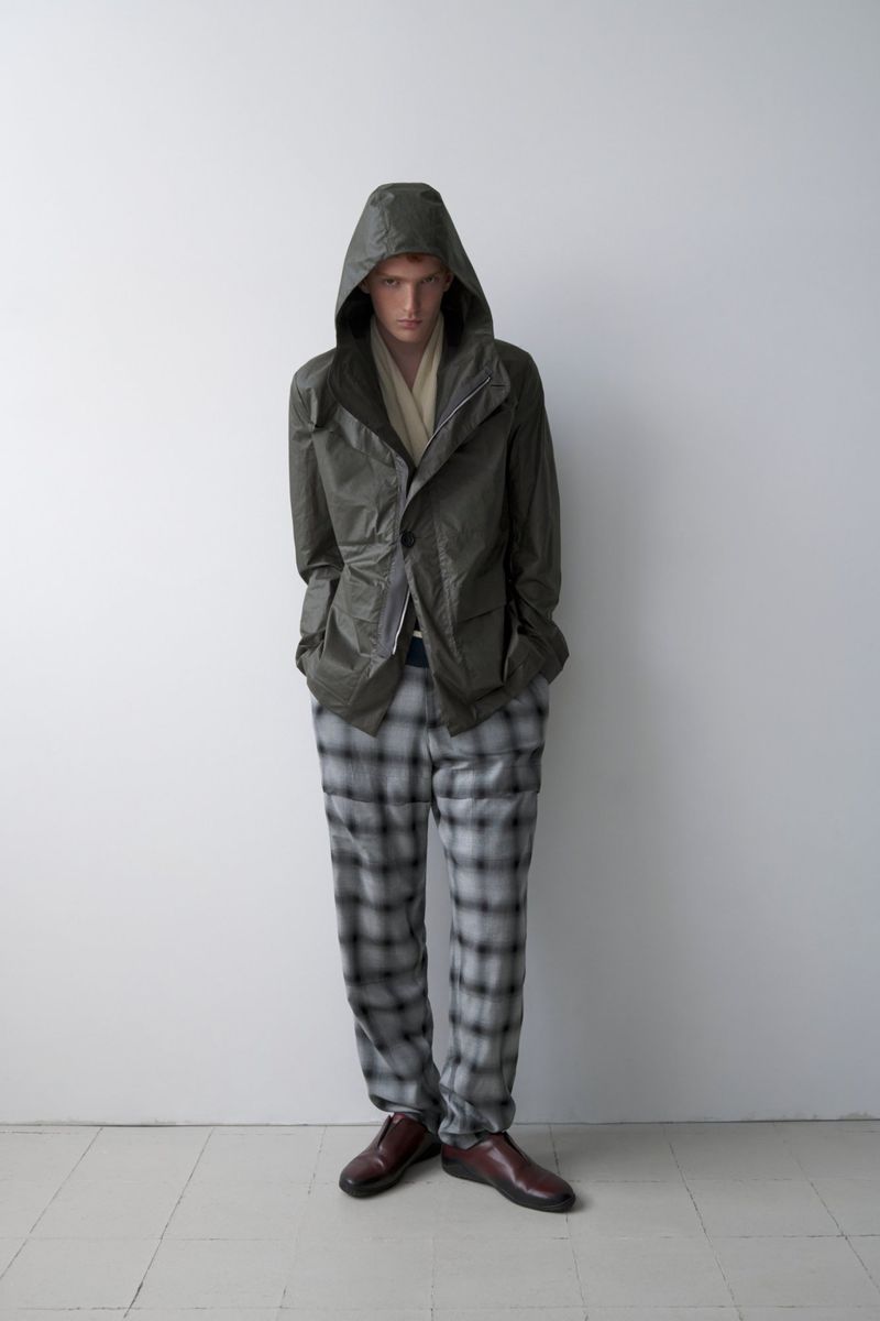 Model Jesse Sachet-Dufraisse wearing an olive hooded jacket and grey plaid trousers against a neutral wall for Derrick S/S 26.