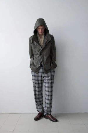 Model Jesse Sachet-Dufraisse wearing an olive hooded jacket and grey plaid trousers against a neutral wall for Derrick S/S 26.