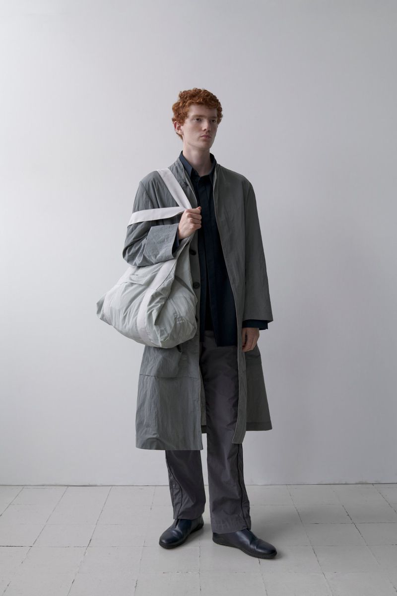 Man in overcoat and pants holding a large bag in a minimalist studio setting with soft lighting and neutral color palette fashion shoot.
