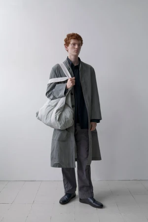 Man in overcoat and pants holding a large bag in a minimalist studio setting with soft lighting and neutral color palette fashion shoot.