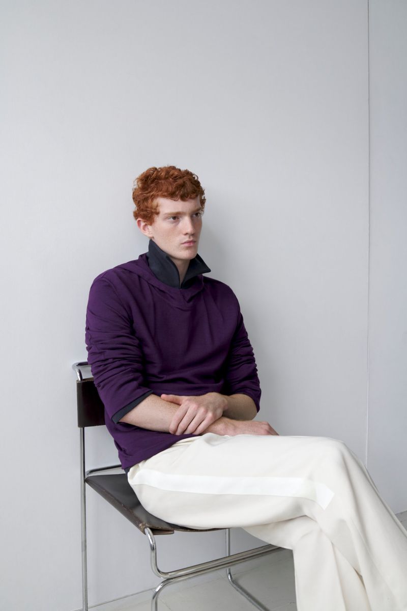 A red-haired adult male model sits on a black chair against a white wall wearing a purple sweater and white pants in a studio setting.
