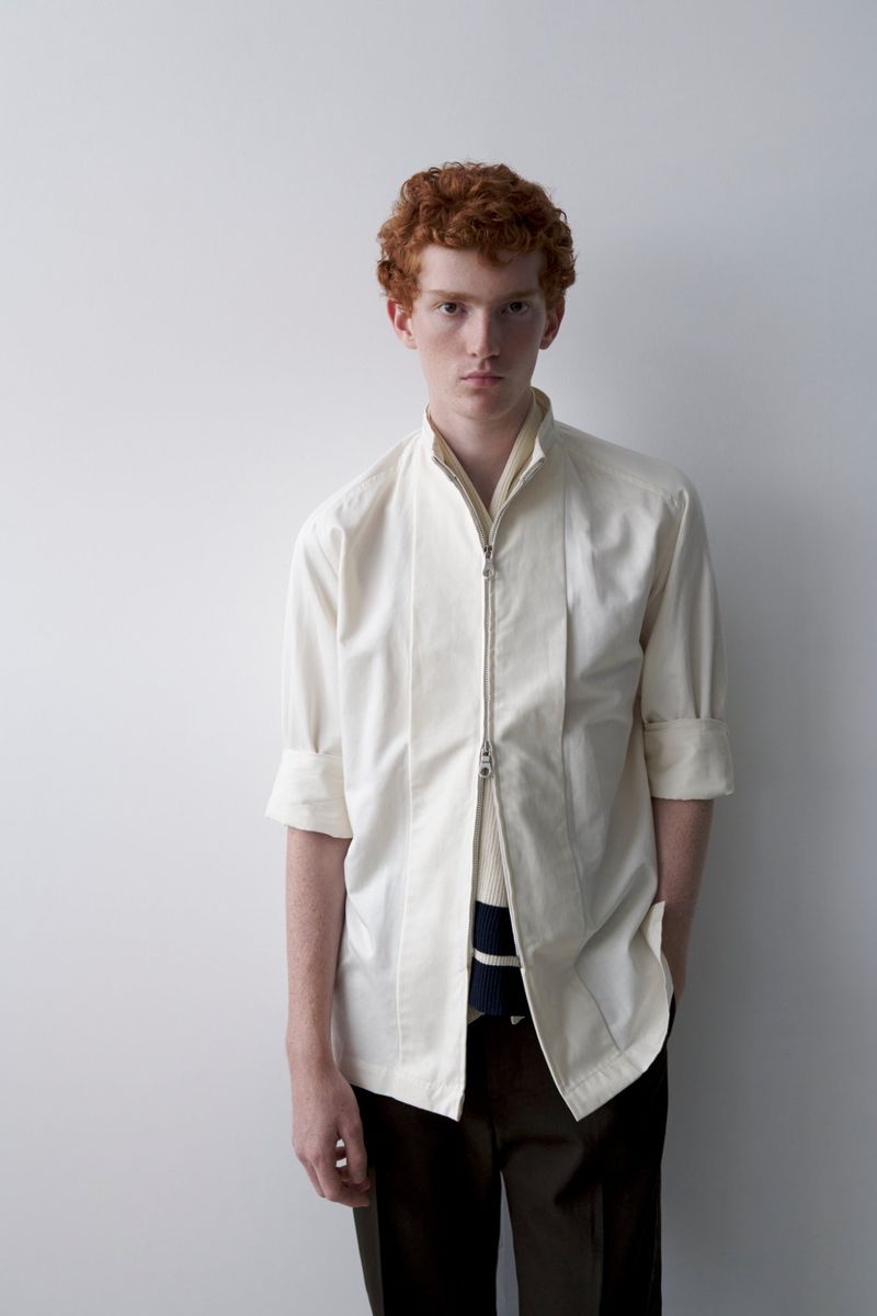 Adult male with red hair posing in a photo studio, wearing a collared off-white shirt with dark pants, against a solid white backdrop.