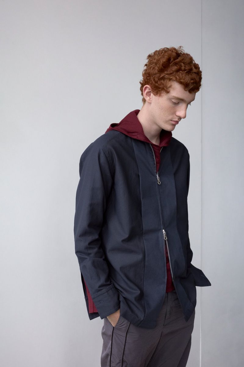 Male fashion model in casual clothing, including a blue zip jacket, maroon hoodie, and grey pants, poses in a neutral studio setting.