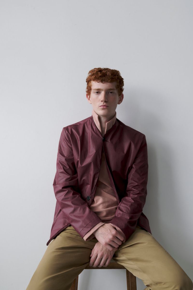 Portrait of a young adult man with red hair and a neutral expression wearing a maroon jacket and tan pants, seated indoors against a plain wall.