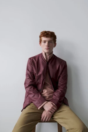 Portrait of a young adult man with red hair and a neutral expression wearing a maroon jacket and tan pants, seated indoors against a plain wall.