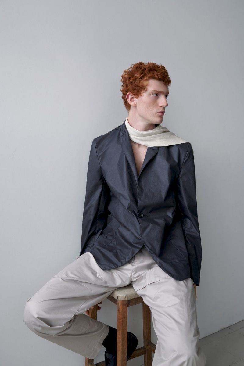 A red-haired person in blazer and trousers poses while seated on stool in an eye-level portrait on a gray studio backdrop.