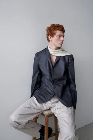 A red-haired person in blazer and trousers poses while seated on stool in an eye-level portrait on a gray studio backdrop.