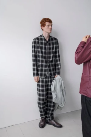 Man in a black and white plaid suit stands in front of a white wall, holding a light gray jacket, with another figure to his right.