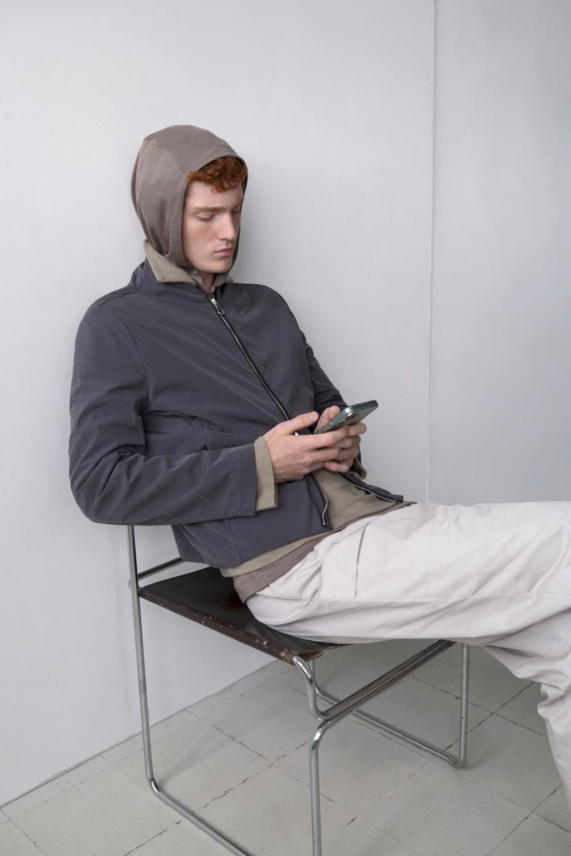 Model Jesse Sachet-Dufraisse seated on a metal frame chair looking at a phone in a grey zip jacket and off-white trousers.